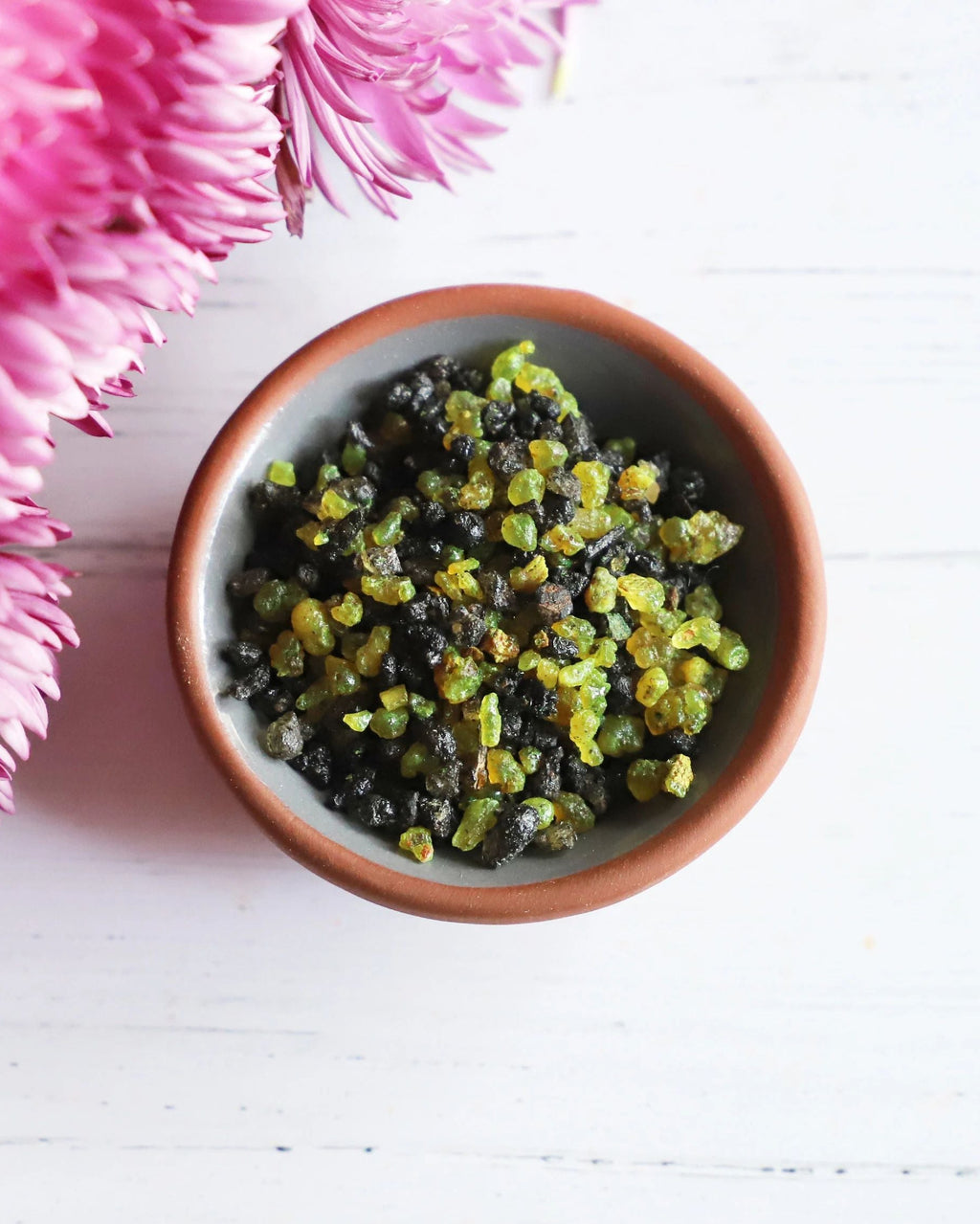 Green and black resin incense in a bowl on a white background with pink flowers next it.