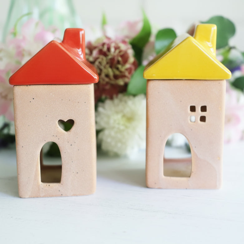 Two small house-shaped essential oil diffusers with colorful roofs on a light surface with flowers in the background.