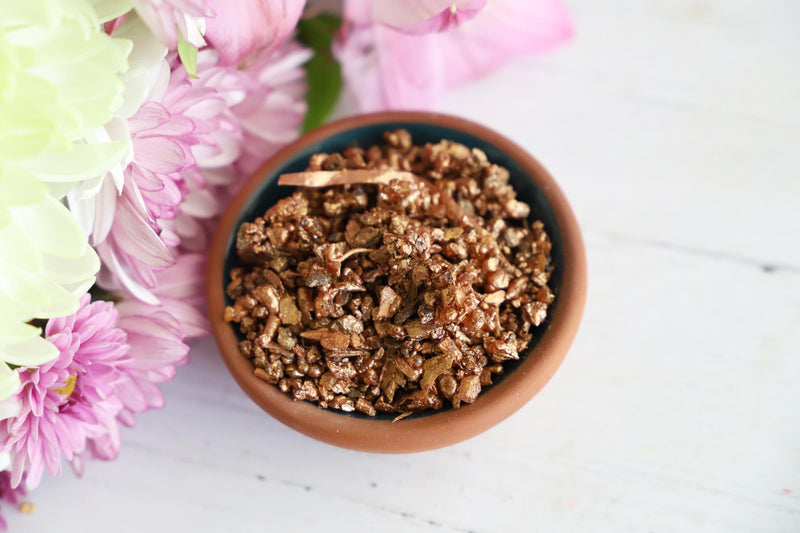 Close-up of copper colored incense granules in a bowl on a wooden white background with fresh flowers next to it.
