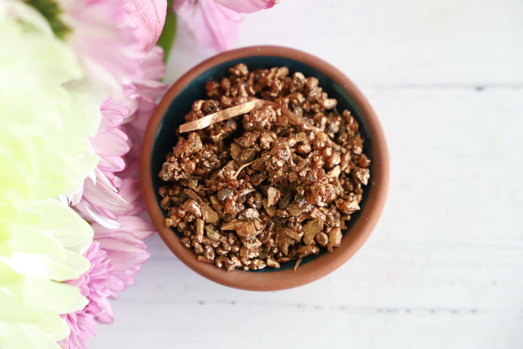 Copper colored incense granules in a bowl on a wooden white background with fresh flowers next to it.