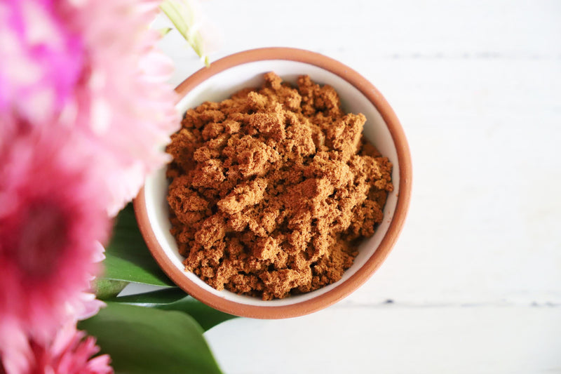 Brown incense powder in a small dish on a white background with colorful fresh flowers on the side.