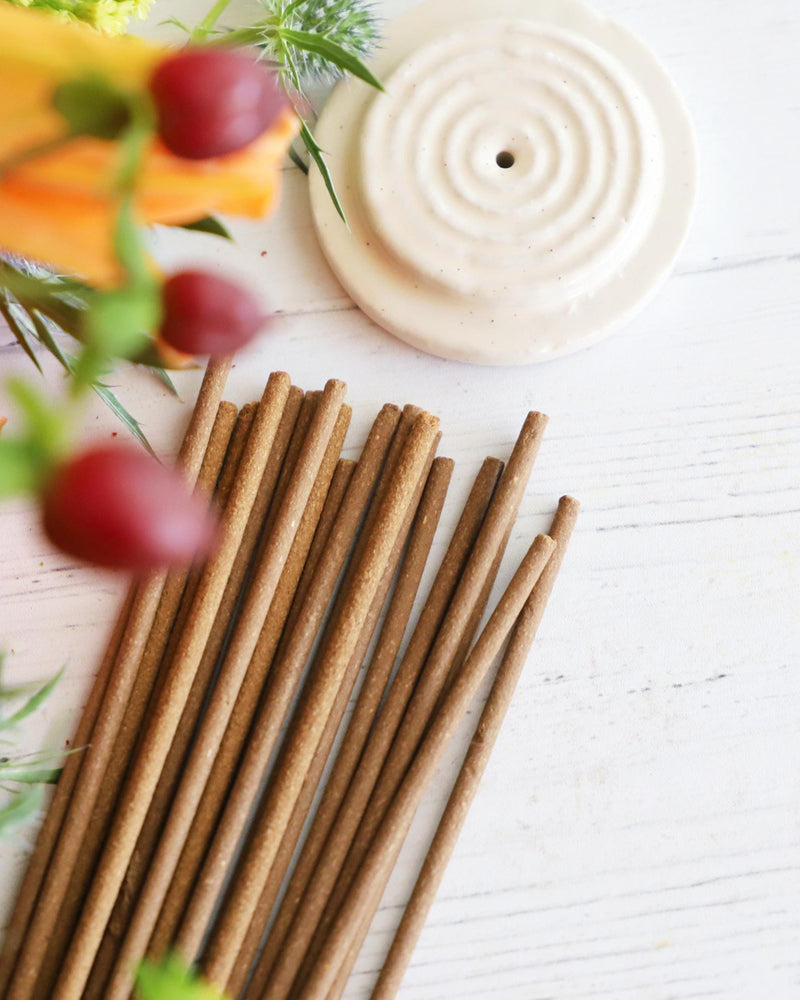 Incense sticks with round white incense holder on white background with colorful flowers next to them.