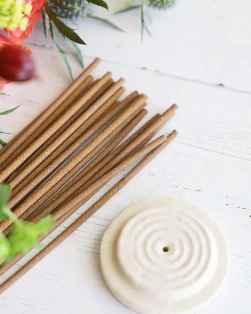 Incense sticks with round white incense holder on white background with colorful flowers next to them.
