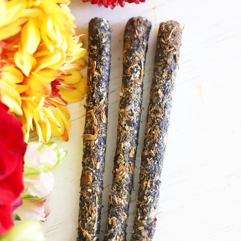 Three incense sticks next to green, yellow, white and red flowers on a white background.