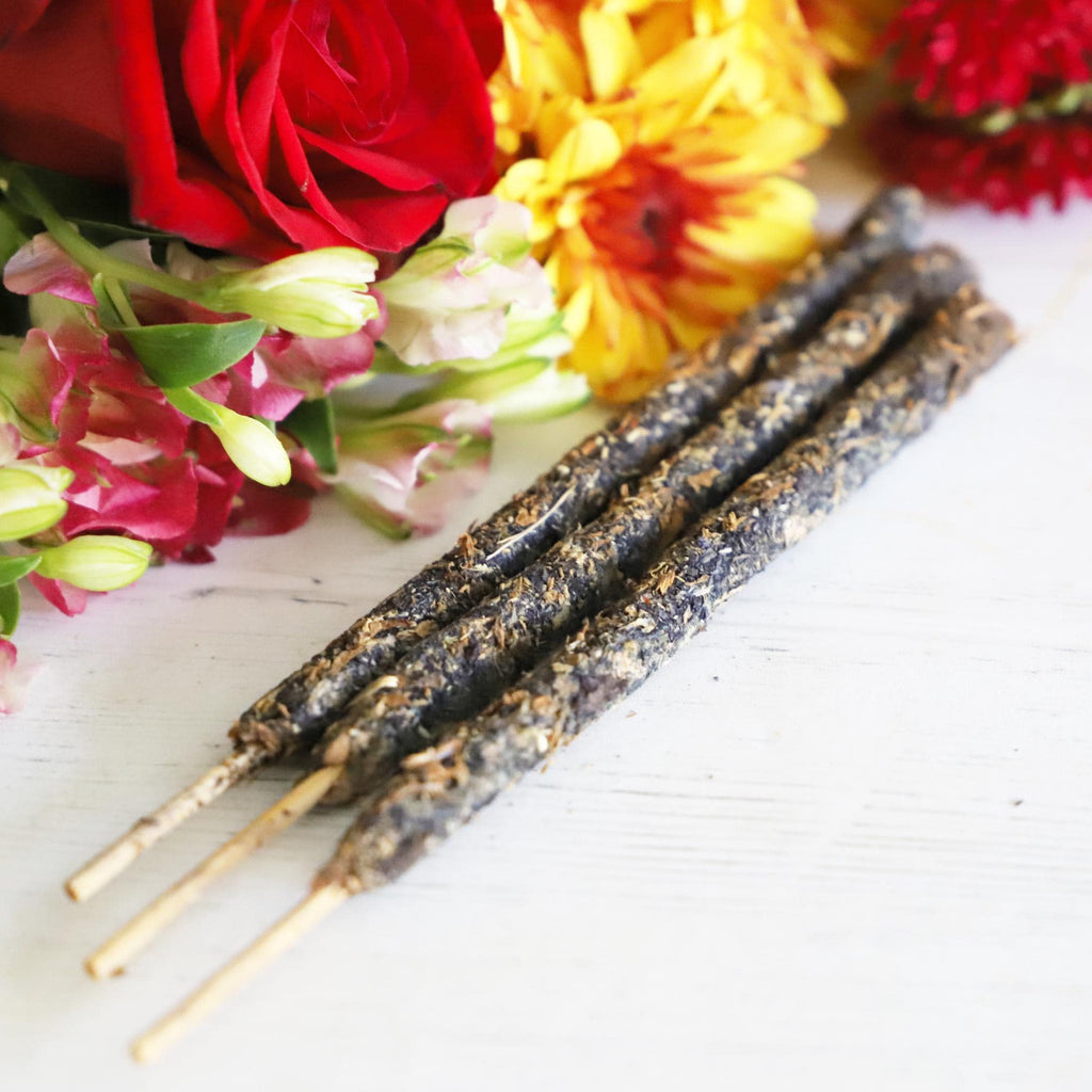 Three incense sticks next to green, yellow, white and red flowers on a white background.