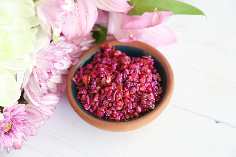 Small bowl of pink resin grains on a white surface with pink flowers in the background