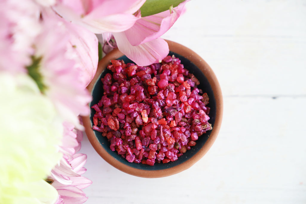 Small bowl of pink resin grains on a white surface with pink flowers in the background