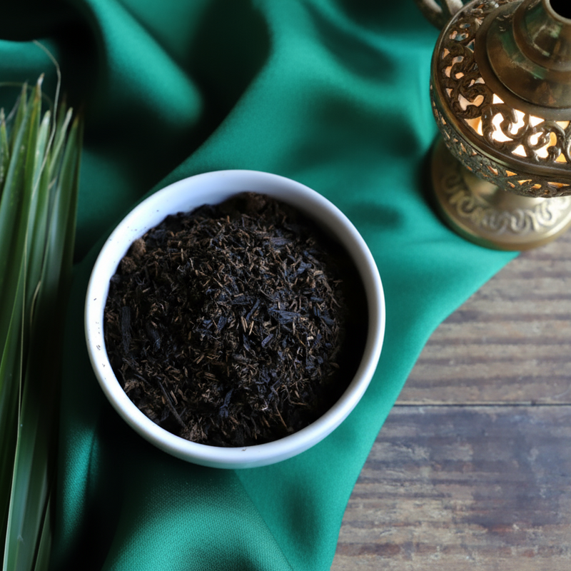 Black incense chips in a white bowl on a green scarf on a wooden table with greenery and a brass lantern
