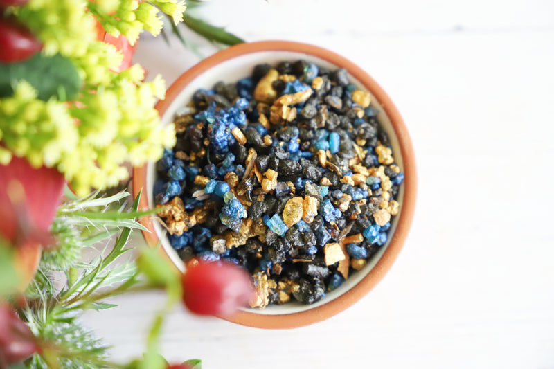 Colorful resin incense grains in a bowl on a white background with fresh red and green flowers next to it.
