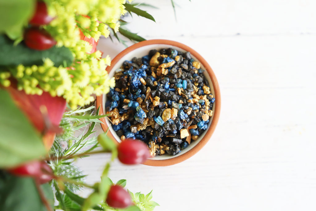 Colorful resin incense grains in a bowl on a white background with fresh red and green flowers next to it.